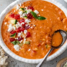 A bowl of gazpacho topped with diced tomatoes, cucumber, cheese, and herbs, with a spoon resting inside.