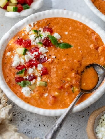 A bowl of gazpacho topped with diced tomatoes, cucumber, cheese, and herbs, with a spoon resting inside.