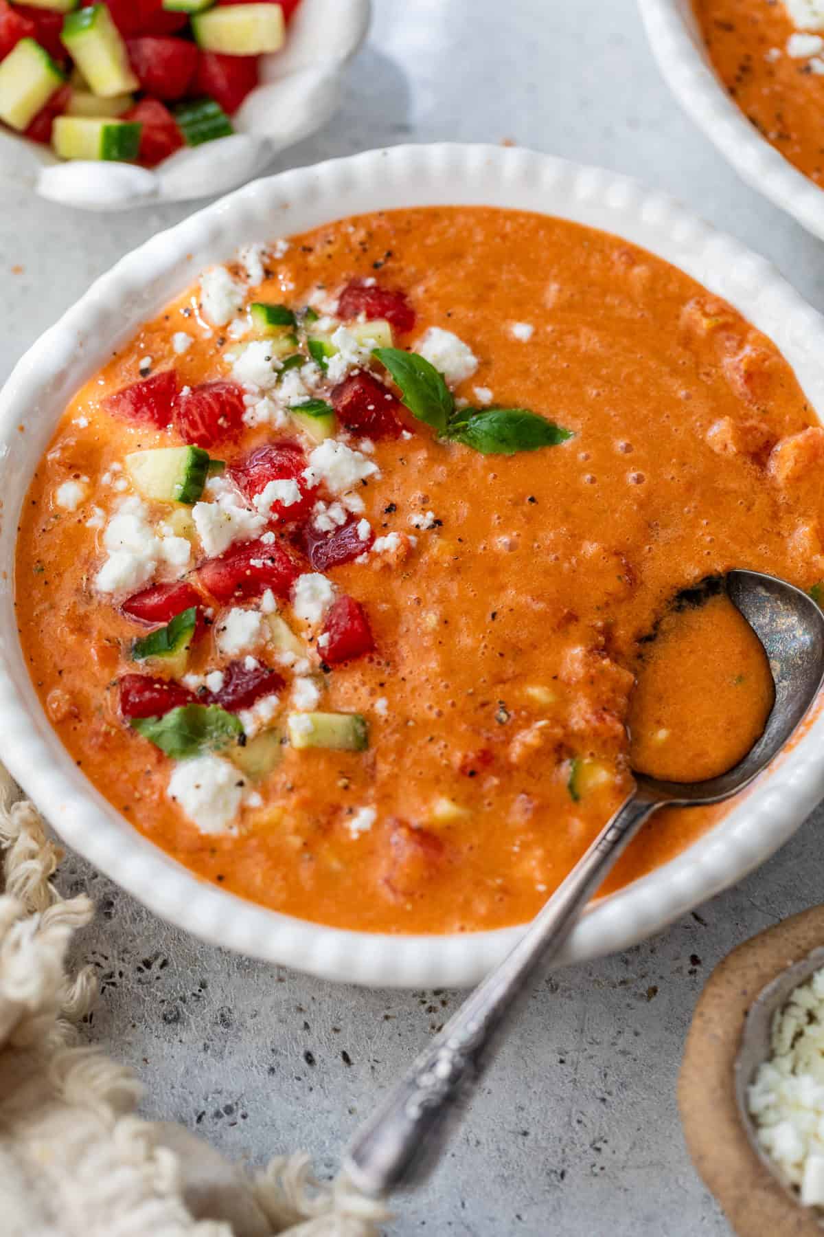 A bowl of gazpacho topped with diced tomatoes, cucumber, cheese, and herbs, with a spoon resting inside.