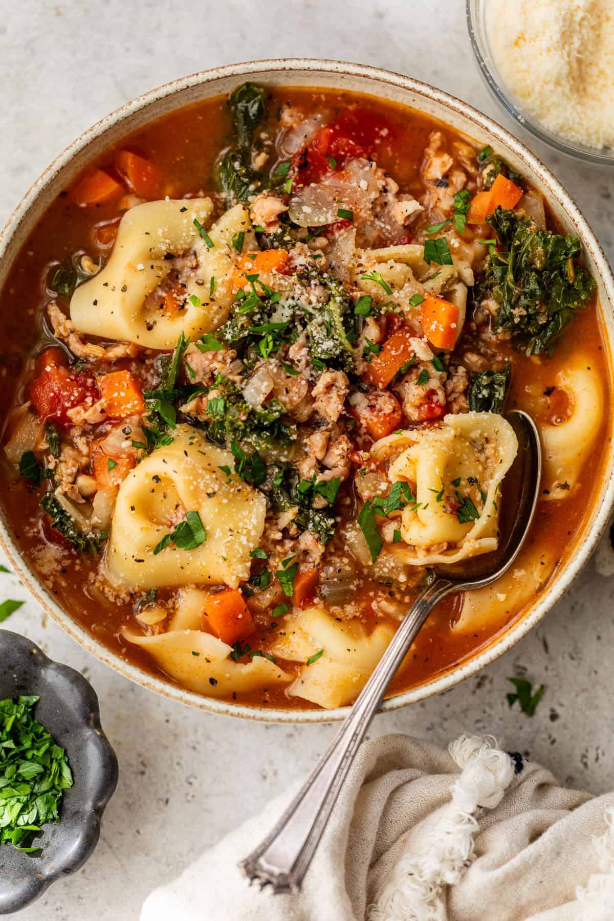 Bowl of crockpot tortellini soup with vegetables, sausage, and herbs, garnished with parsley and grated cheese, with a spoon.