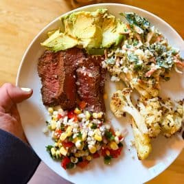 A plate with sliced steak, avocado, corn salad, creamy slaw, and roasted cauliflower, held over a wooden floor.