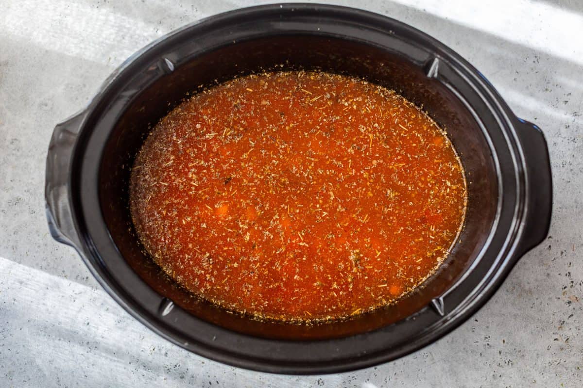 A slow cooker filled with red tomato-based broth sprinkled with herbs, on a light surface.