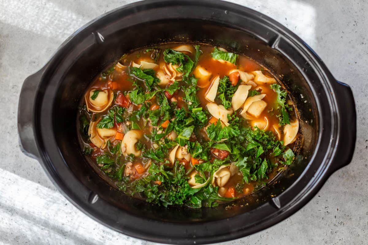 Top view of tortellini soup with kale in a black crockpot on a light gray surface.