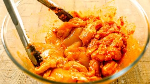 Raw chicken pieces marinated in a harissa marinade in a glass bowl with metal tongs on a wooden surface.