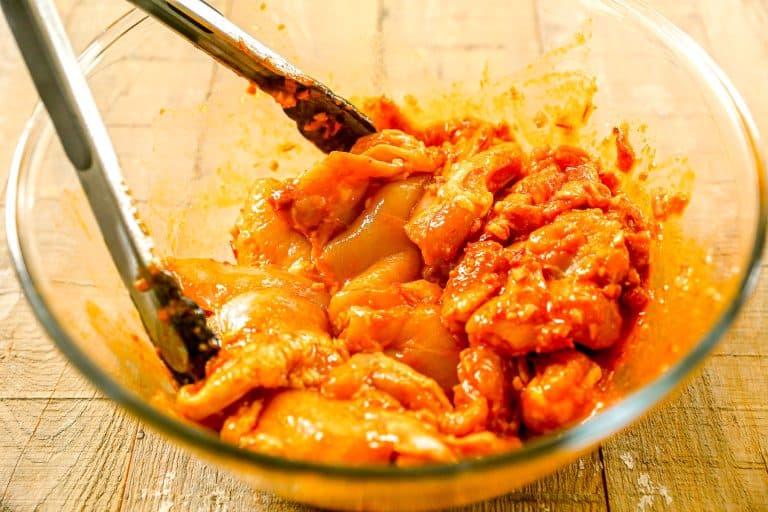 Raw chicken pieces marinated in a harissa marinade in a glass bowl with metal tongs on a wooden surface.