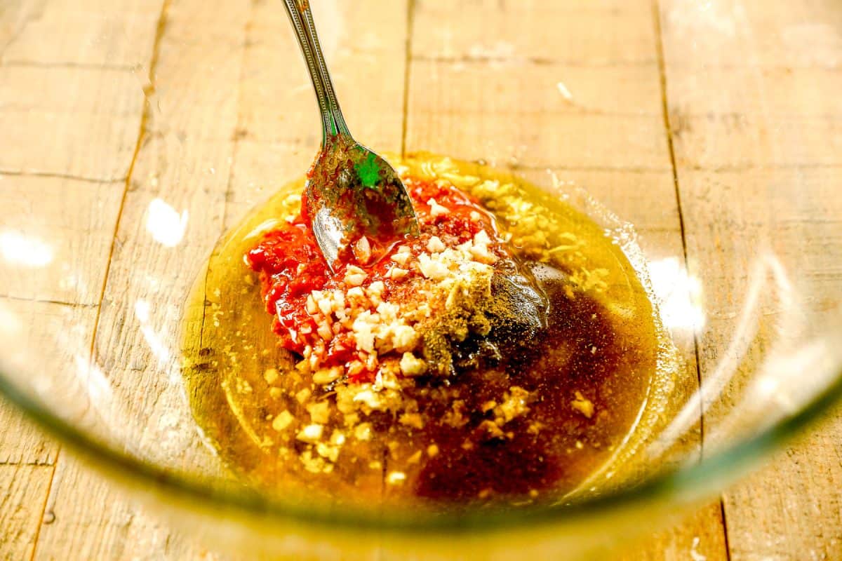 A spoon mixes oil, minced garlic, spices, and harissa in a clear glass bowl on a wooden surface.