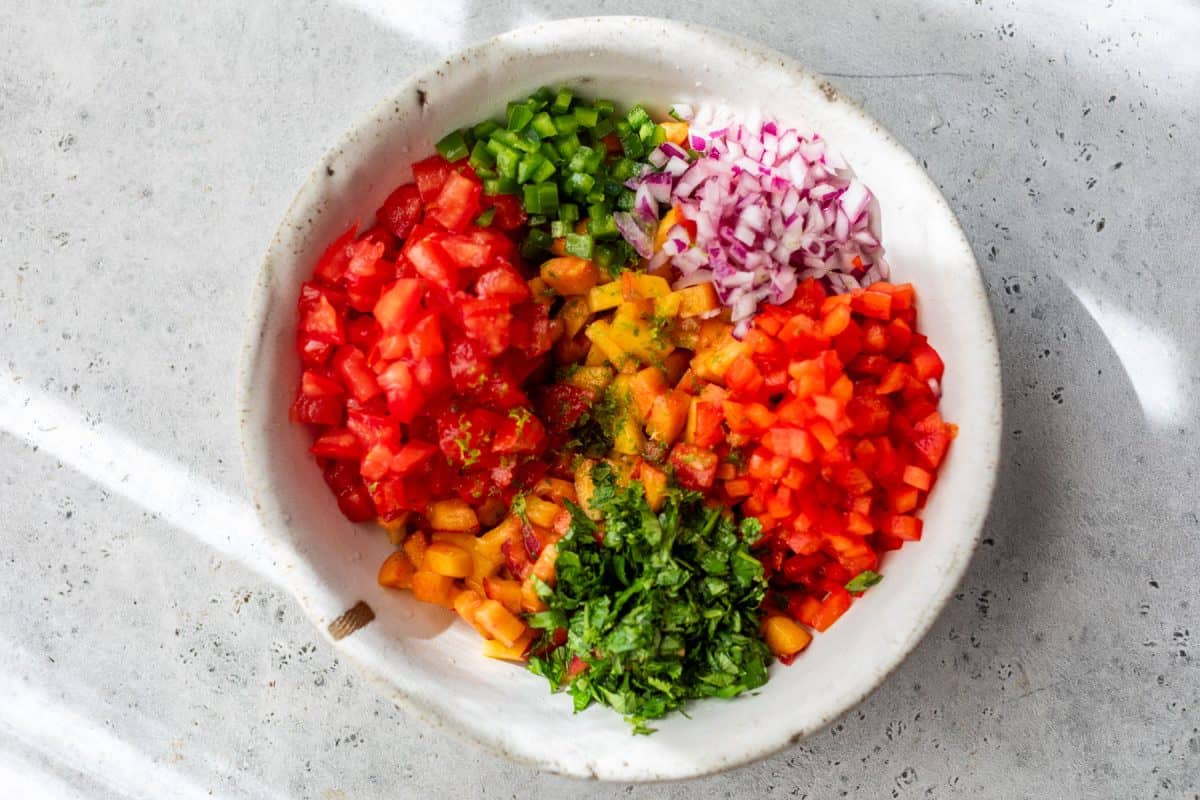 Bowl with chopped tomato, peaches, bell pepper, red onion, jalapeño, and cilantro on a light surface.