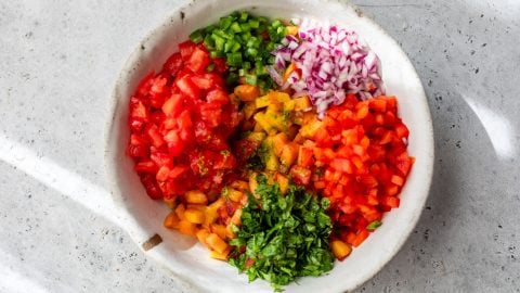 Bowl with chopped tomato, peaches, bell pepper, red onion, jalapeño, and cilantro on a light surface.