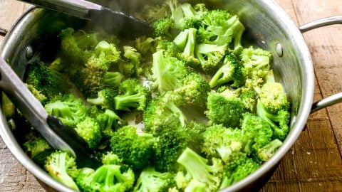 Steamed broccoli florets in a metal pot with black kitchen tongs on a wooden surface.