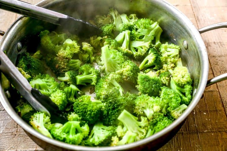 Steamed broccoli florets in a metal pot with black kitchen tongs on a wooden surface.