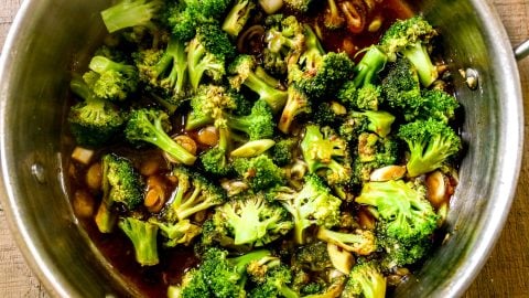 Broccoli florets cooking in a pan with sliced garlic and a dark sauce, seen from above on a wooden surface.