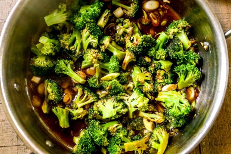 Broccoli florets cooking in a pan with sliced garlic and a dark sauce, seen from above on a wooden surface.