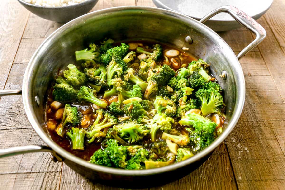 A stainless steel pan filled with broccoli florets in a brown sauce, sitting on a wooden table.