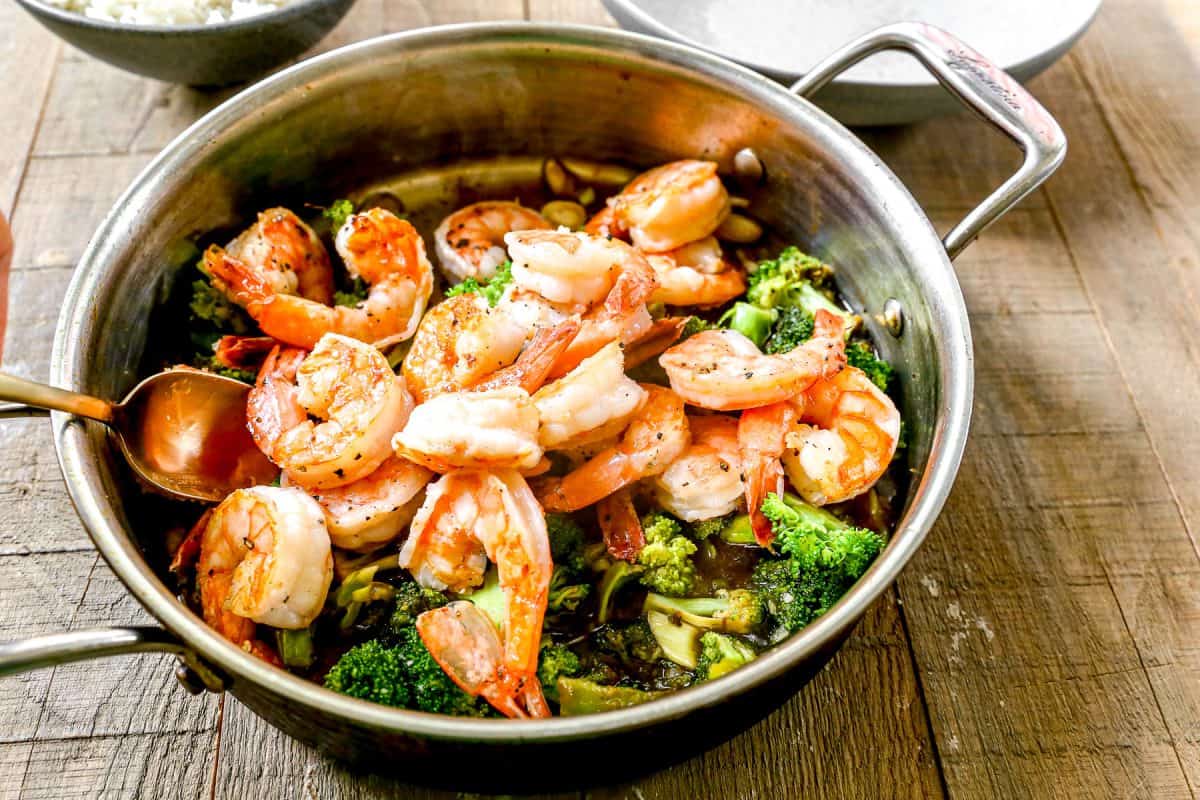 Cooked shrimp and broccoli in a pan with sauce, placed on a wooden table, with a spoon and rice nearby.