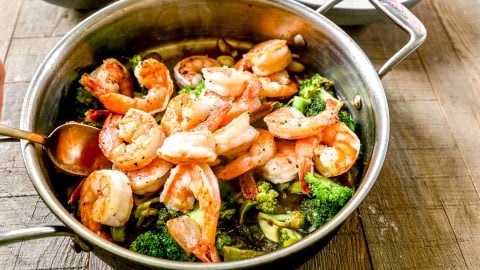 Cooked shrimp and broccoli in a pan with sauce, placed on a wooden table, with a spoon and rice nearby.