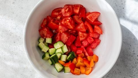 Chopped tomatoes, cucumber, and bell pepper in a white bowl on a light gray surface.