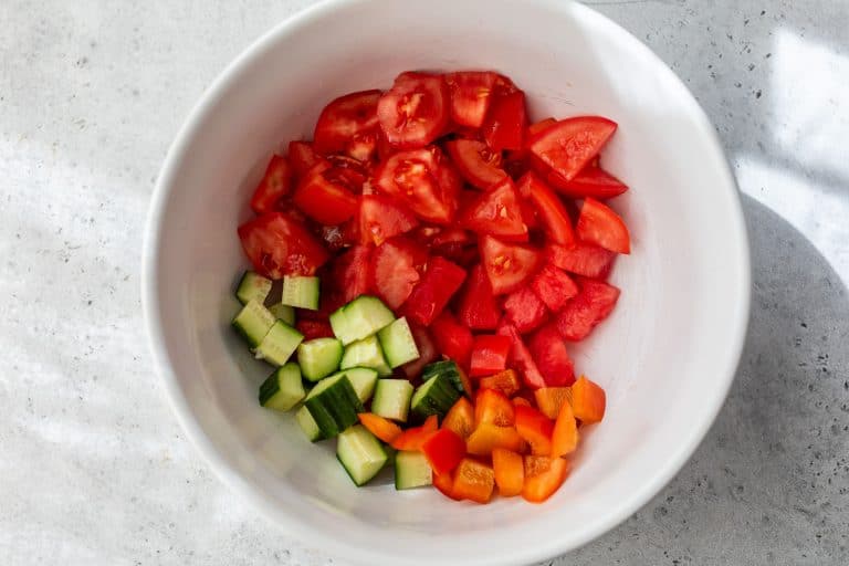 Chopped tomatoes, cucumber, and bell pepper in a white bowl on a light gray surface.
