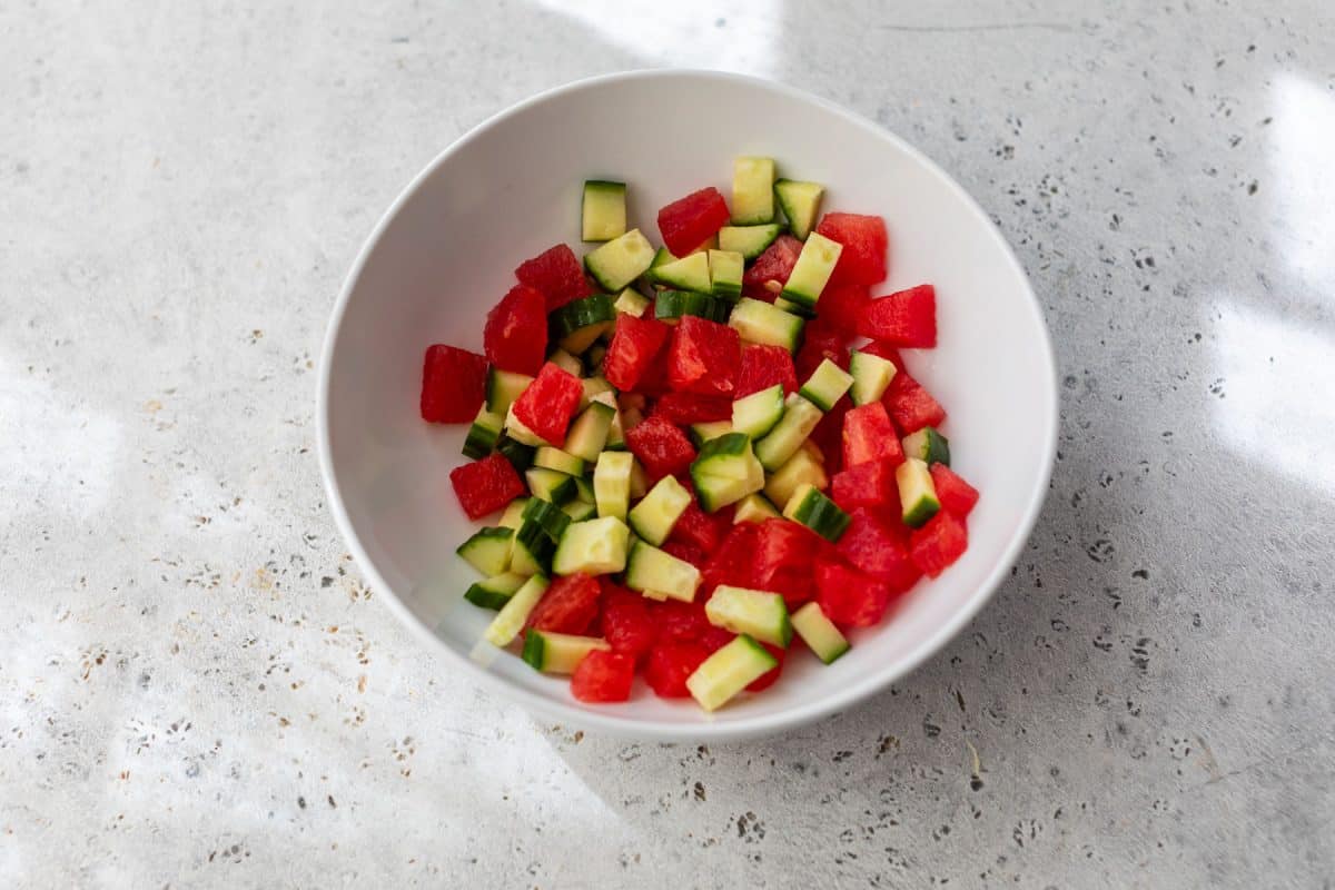 A white bowl filled with diced watermelon, avocado, and cucumber on a light stone surface.