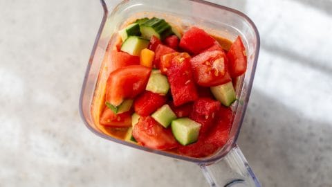Chopped tomatoes and cucumbers in a blender pitcher, ready to be blended, on a light surface.