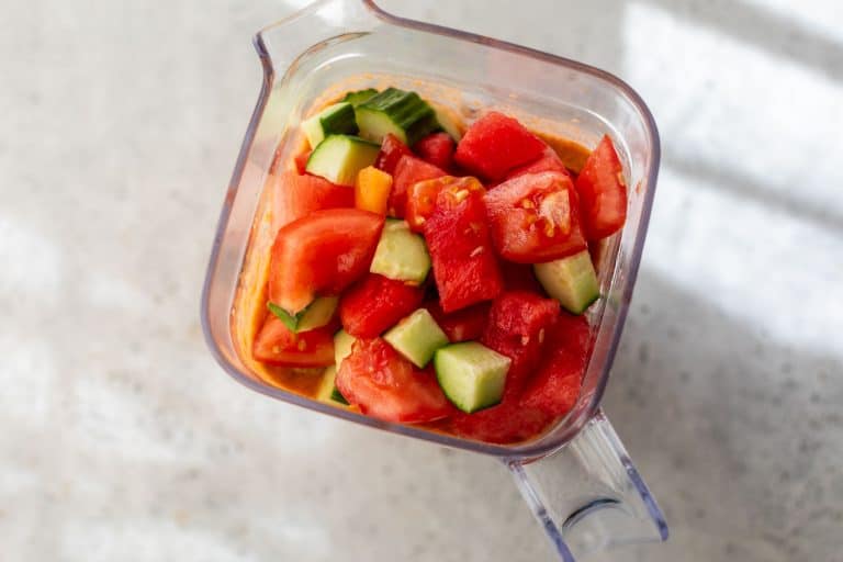 Chopped tomatoes and cucumbers in a blender pitcher, ready to be blended, on a light surface.