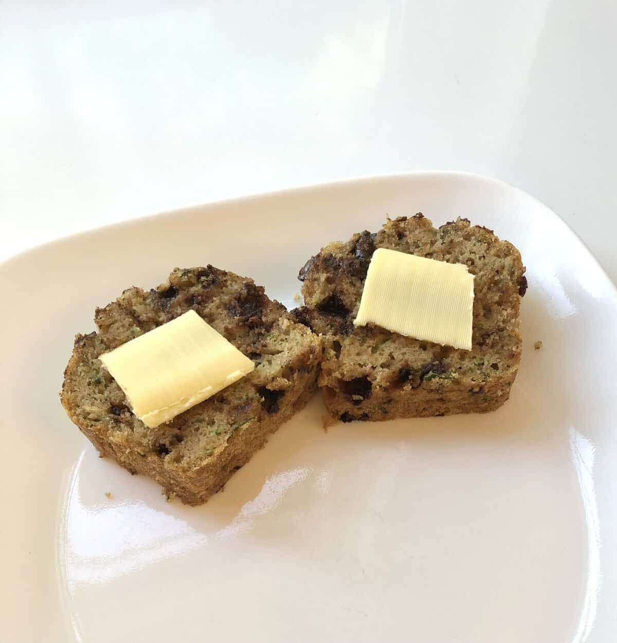 Two slices of zucchini bread with chocolate chips, each topped with a pat of butter, on a white plate.