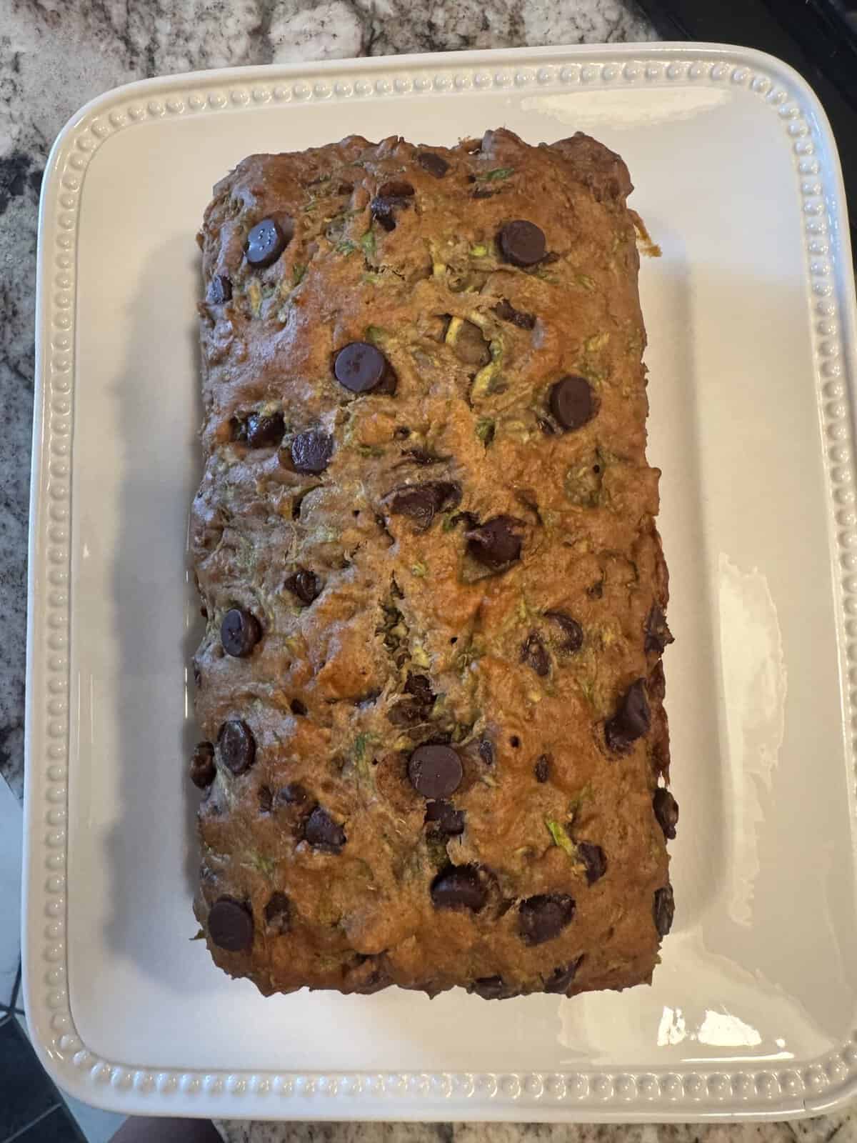 A loaf of zucchini bread with chocolate chips sits on a white square plate with a beaded edge on a countertop.