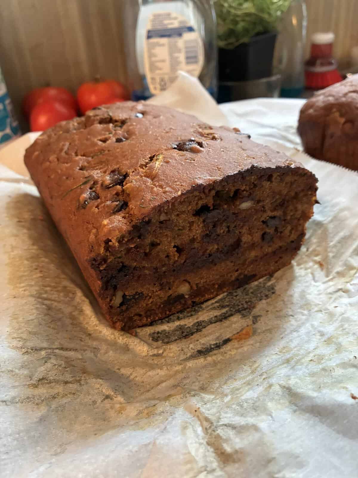 A loaf of banana bread with nuts and chocolate chips, partially sliced, rests on parchment paper in a kitchen setting.