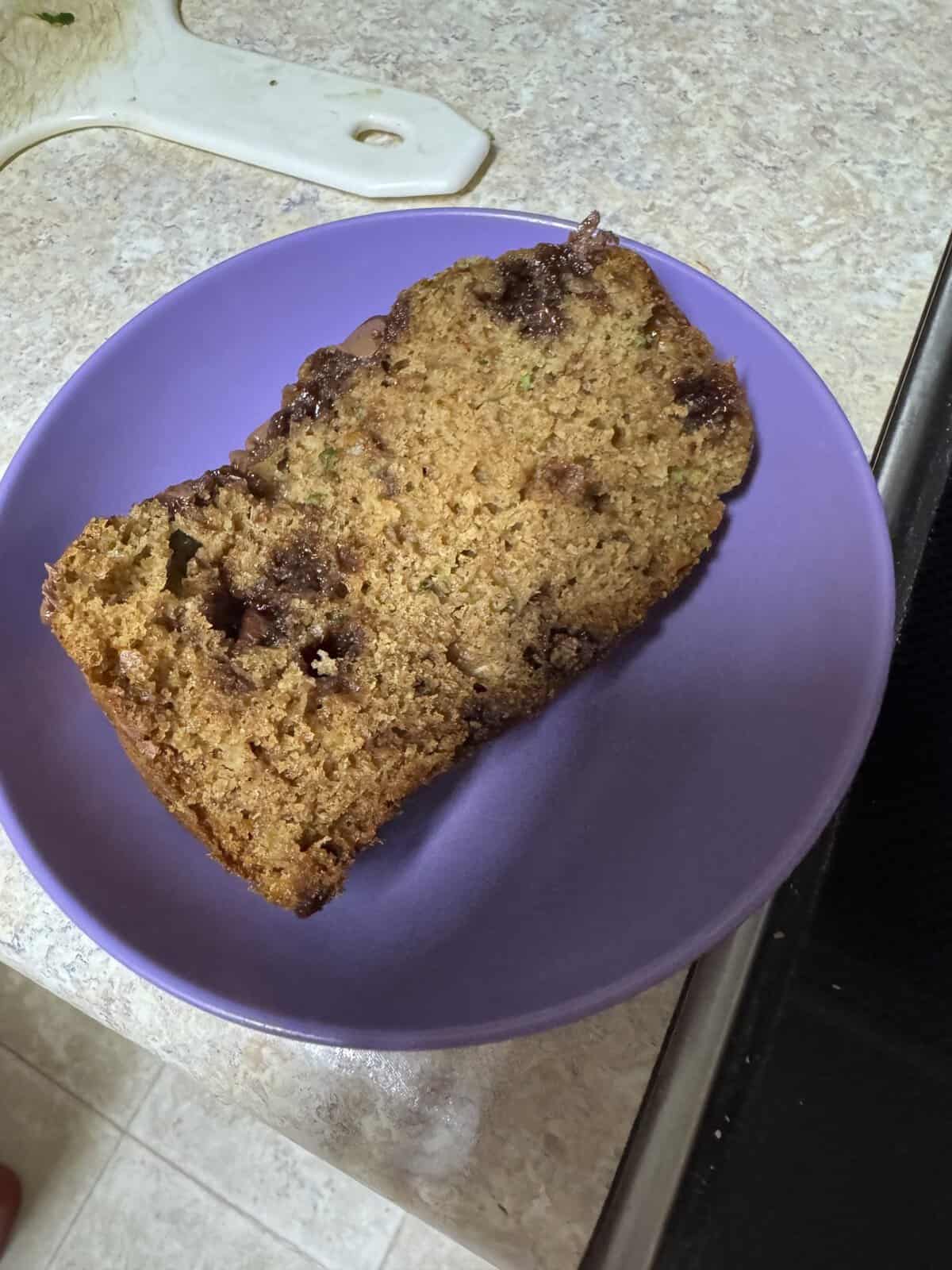 A slice of zucchini bread with chocolate chips on a purple plate, placed on a kitchen counter.