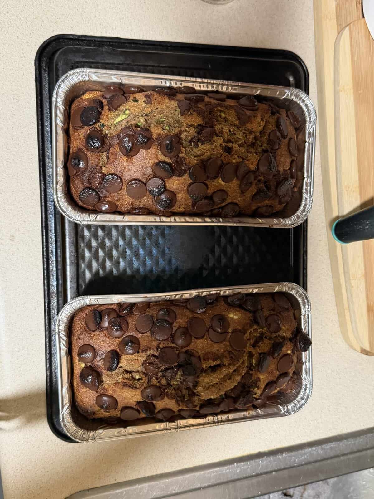 Two loaves of chocolate chip banana bread in foil pans on a baking tray, fresh from the oven.