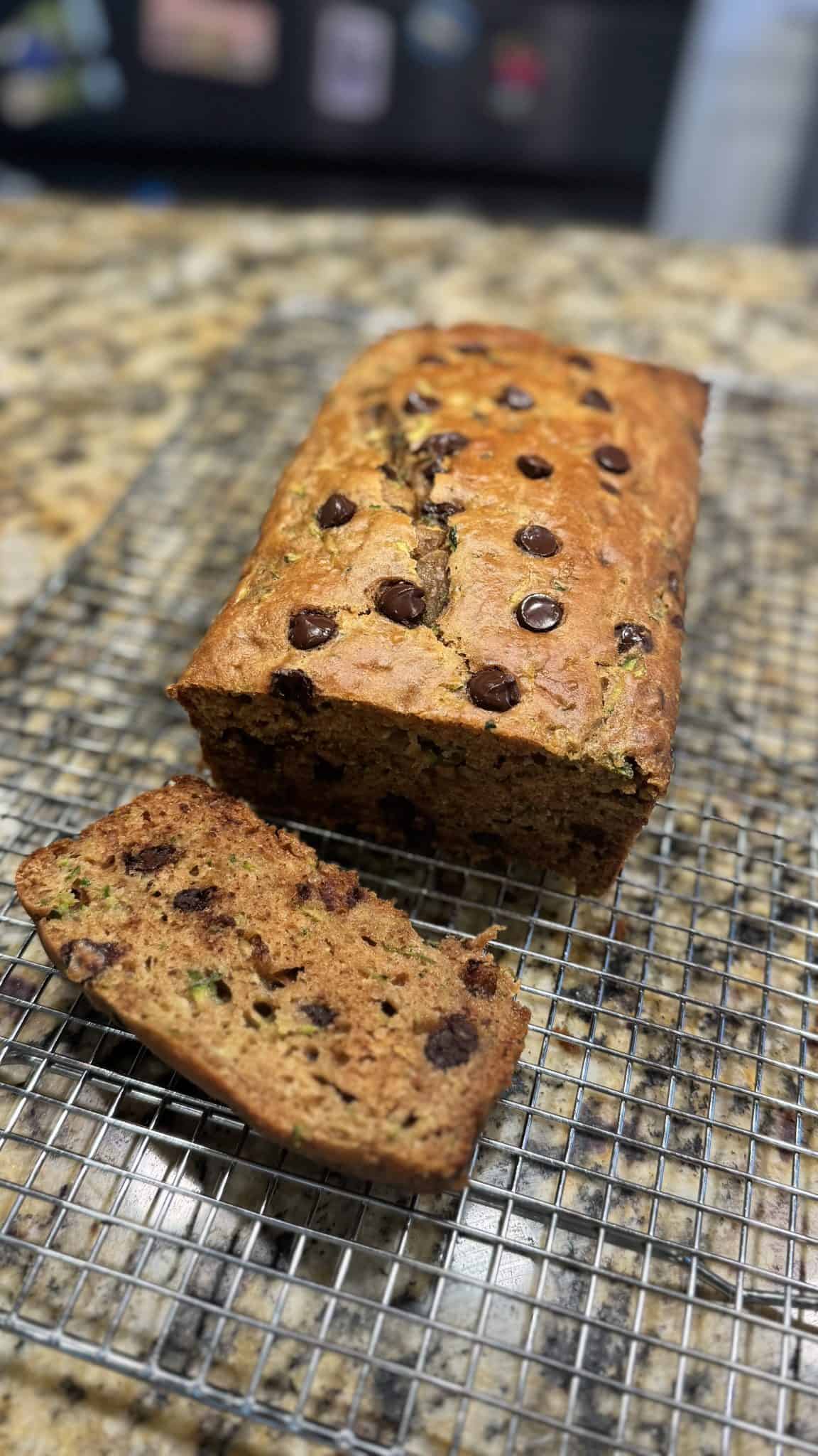 A loaf of chocolate chip zucchini bread with one slice cut, resting on a cooling rack on a granite counter.