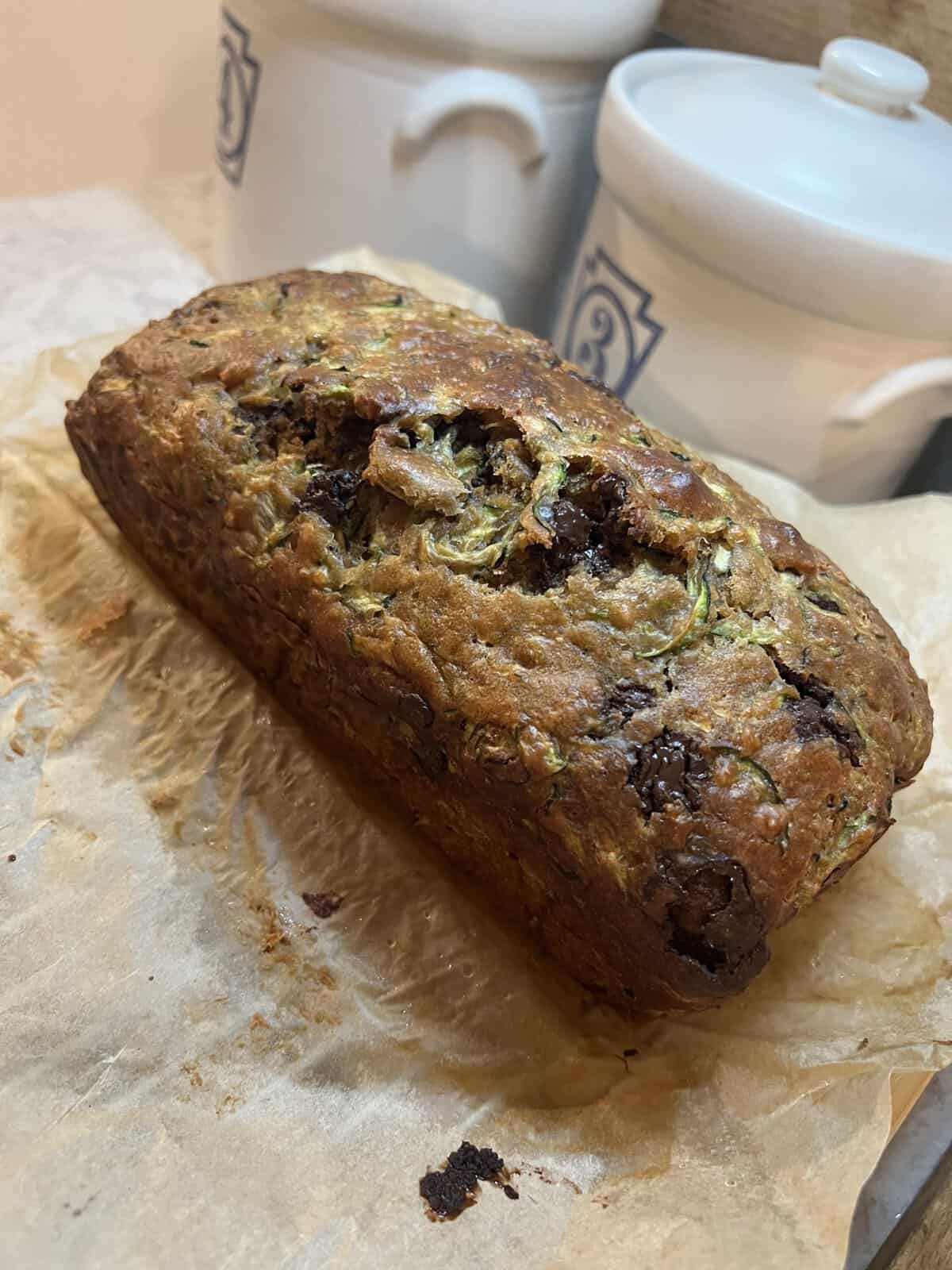 A loaf of homemade zucchini bread with chocolate chips rests on parchment paper near white ceramic canisters.