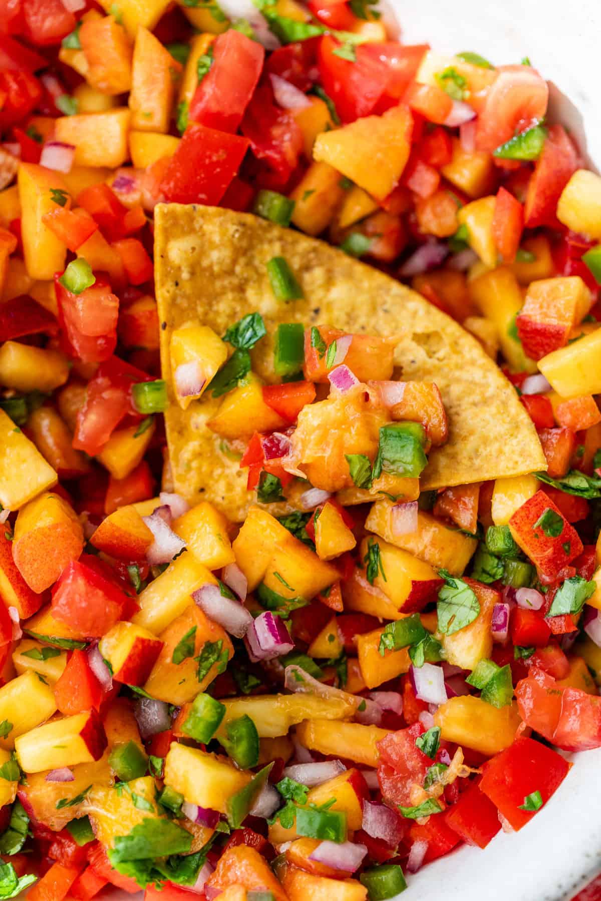 A tortilla chip scoops colorful peach salsa with tomatoes, onions, jalapeños, and cilantro.