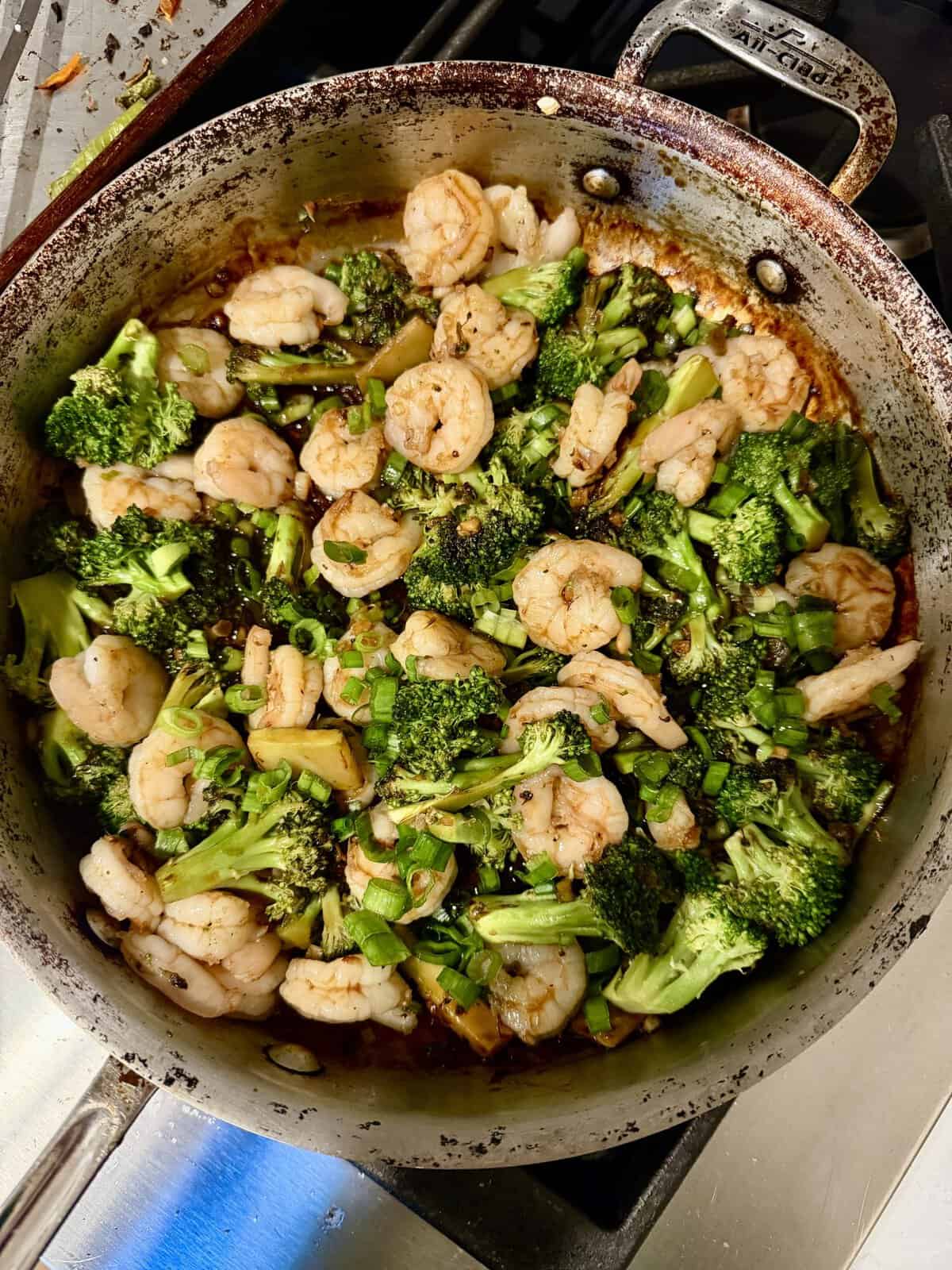 Shrimp and broccoli stir-fry cooking in a metal skillet with green onions and a savory sauce.