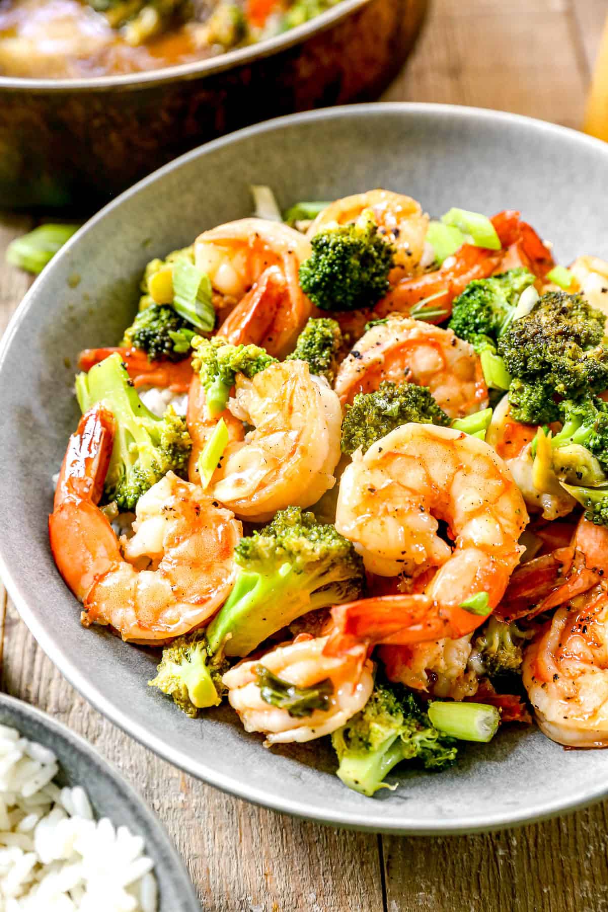 Shrimp and broccoli stir-fry served in a gray bowl, garnished with green onions, next to a dish of white rice.