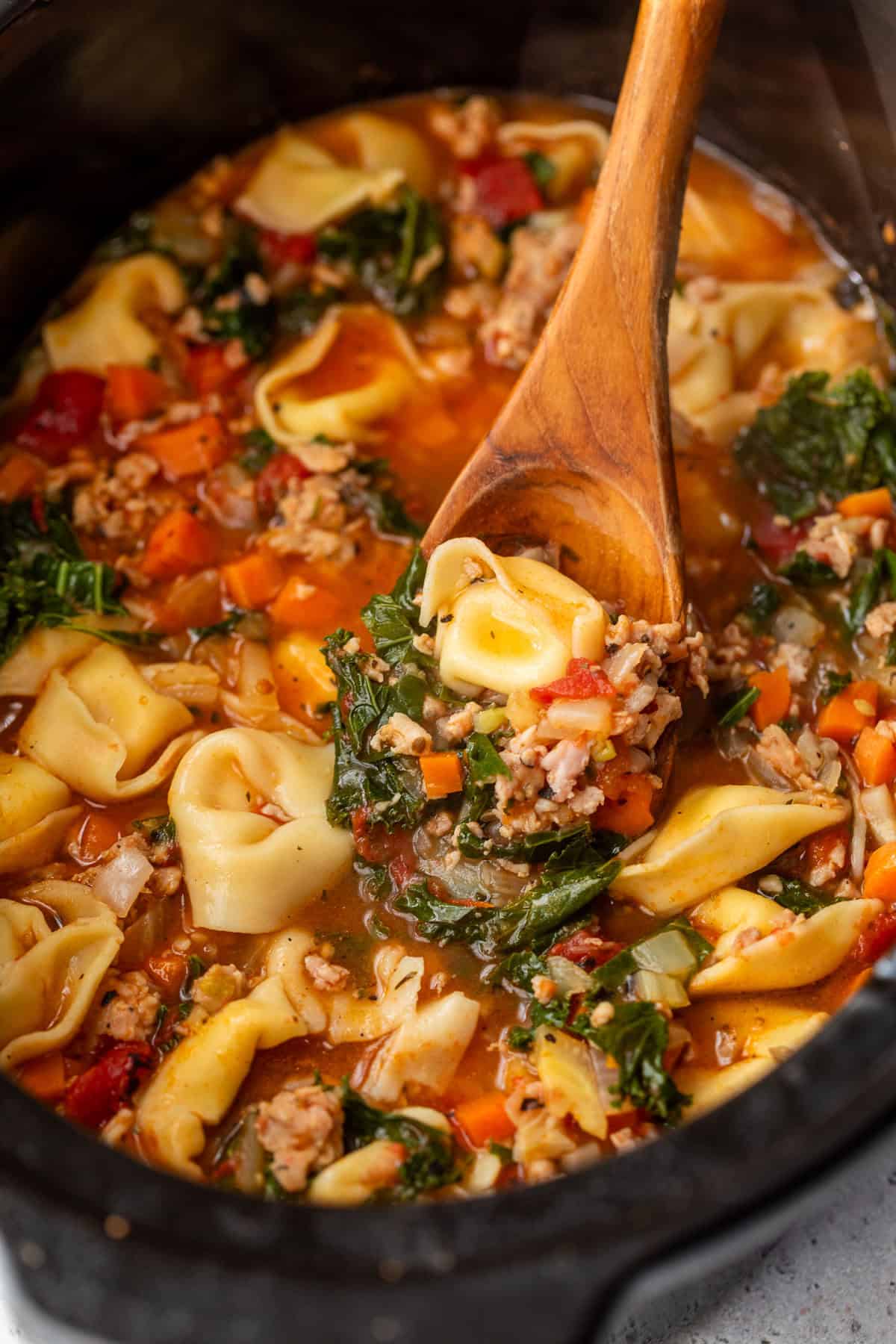 Tortellini soup with sausage, kale, carrots, and tomatoes in a crockpot, stirred with a wooden spoon.