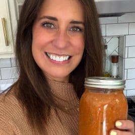 Woman smiling in a kitchen, holding a glass jar filled with homemade tomato sauce.