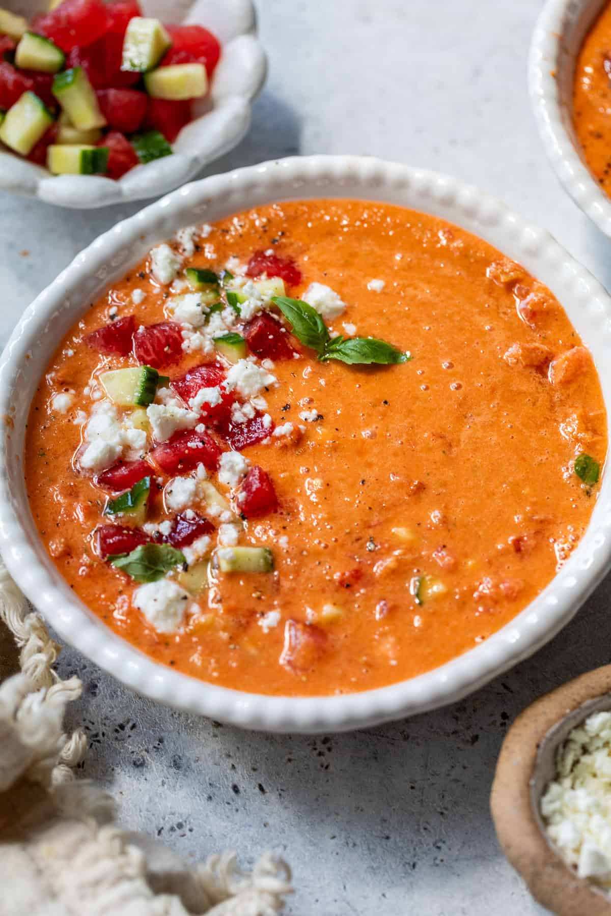 A bowl of creamy watermelon gazpacho soup topped with diced cucumber, tomatoes, feta cheese, and fresh basil leaves.