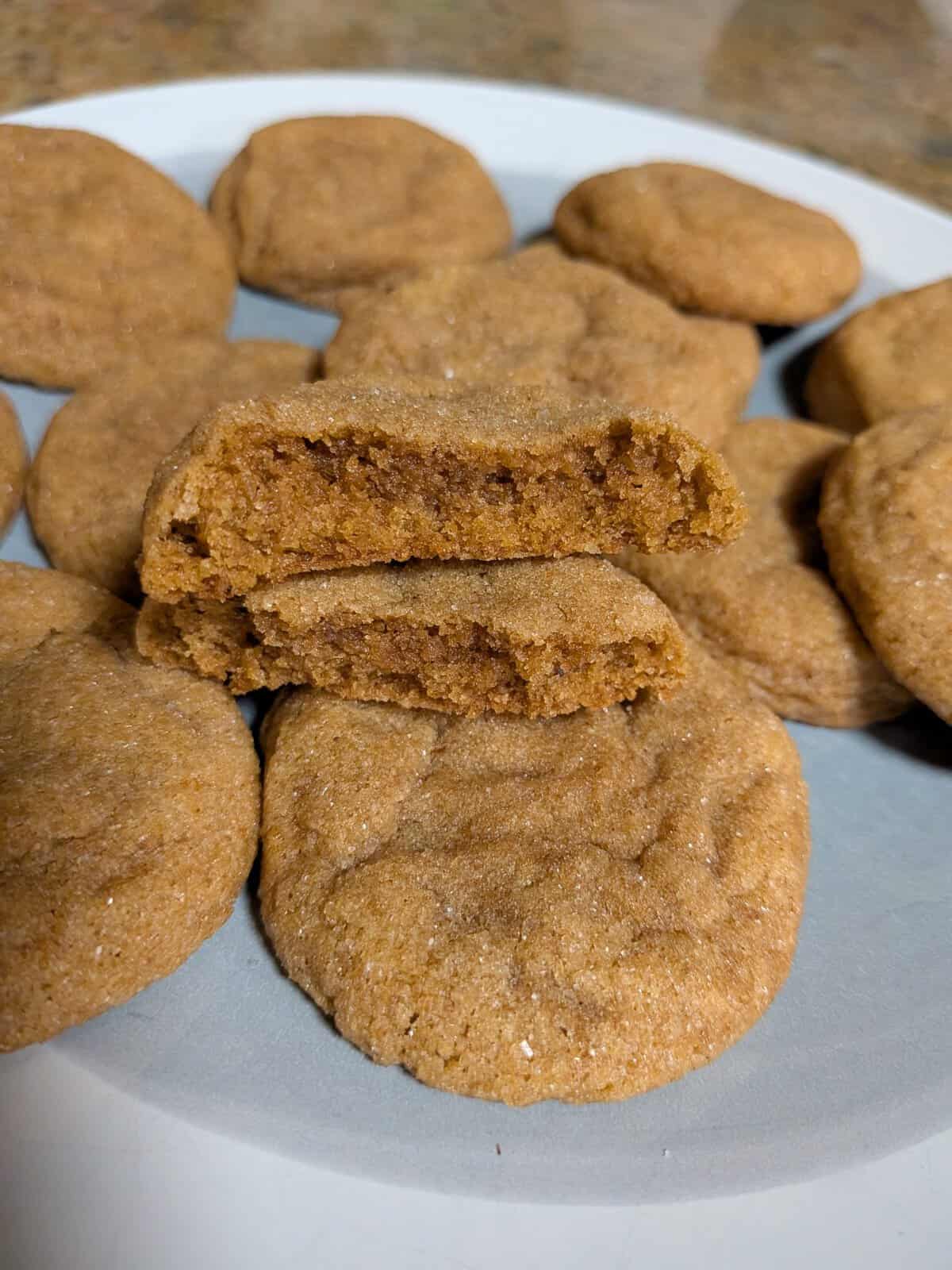A plate of soft, round brown cookies with one cookie broken in half to show its moist inside.