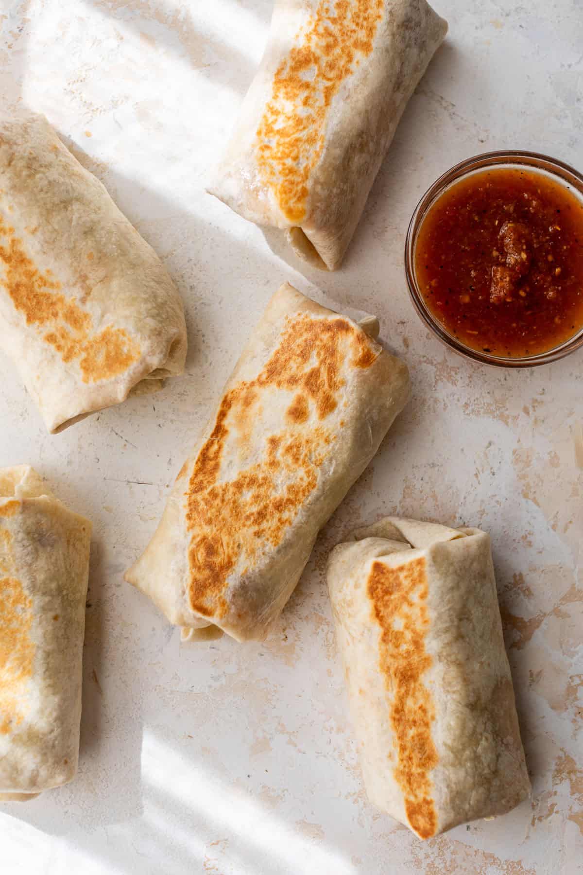 Five browned bean and cheese burritos on a light surface next to a small bowl of red salsa.
