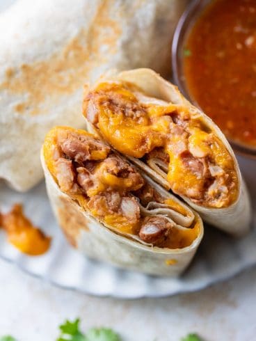 A bean and cheese burrito cut in half, filled with beans and melted cheese, next to a bowl of red salsa and a sprig of cilantro.
