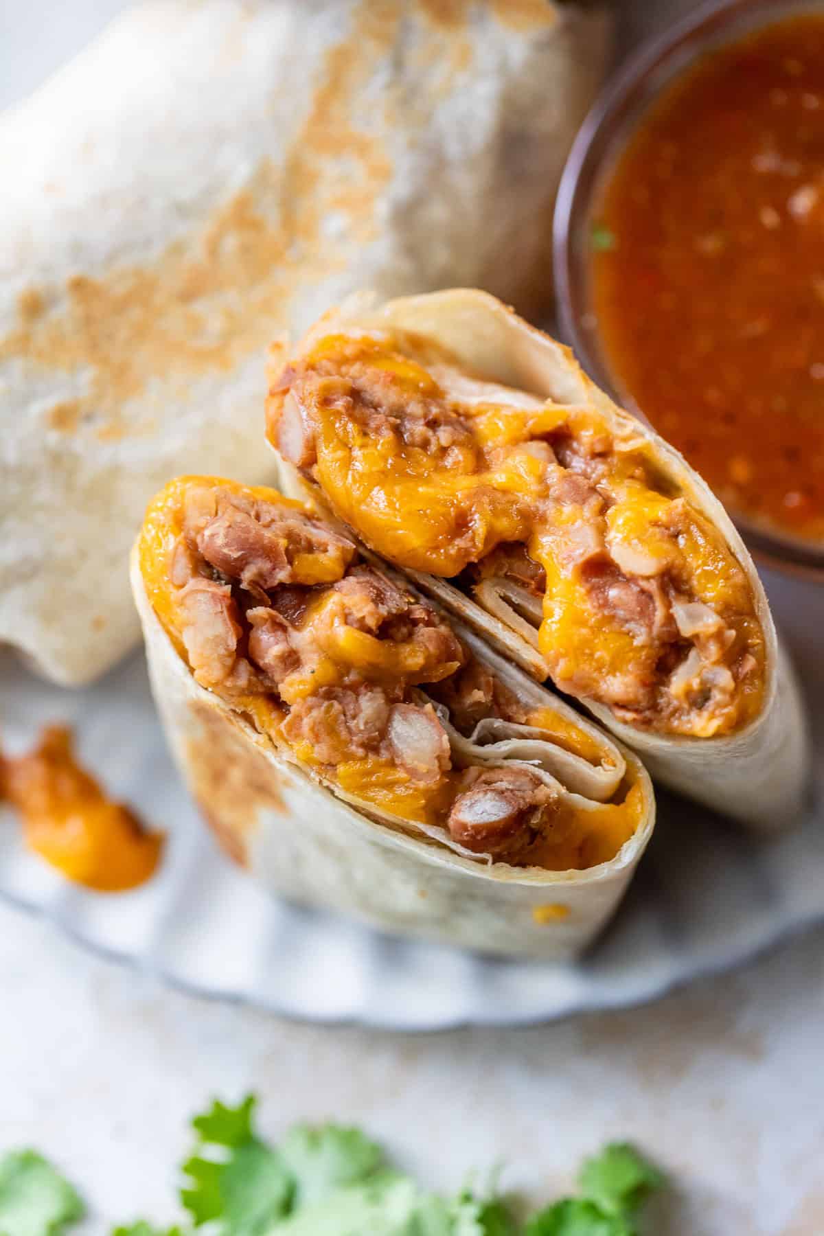 A bean and cheese burrito cut in half, filled with beans and melted cheese, next to a bowl of red salsa and a sprig of cilantro.