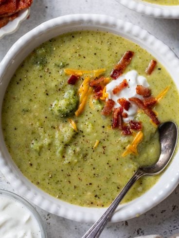 Creamy broccoli potato soup topped with shredded cheese, bacon bits, and a dollop of sour cream in a white bowl with a spoon.