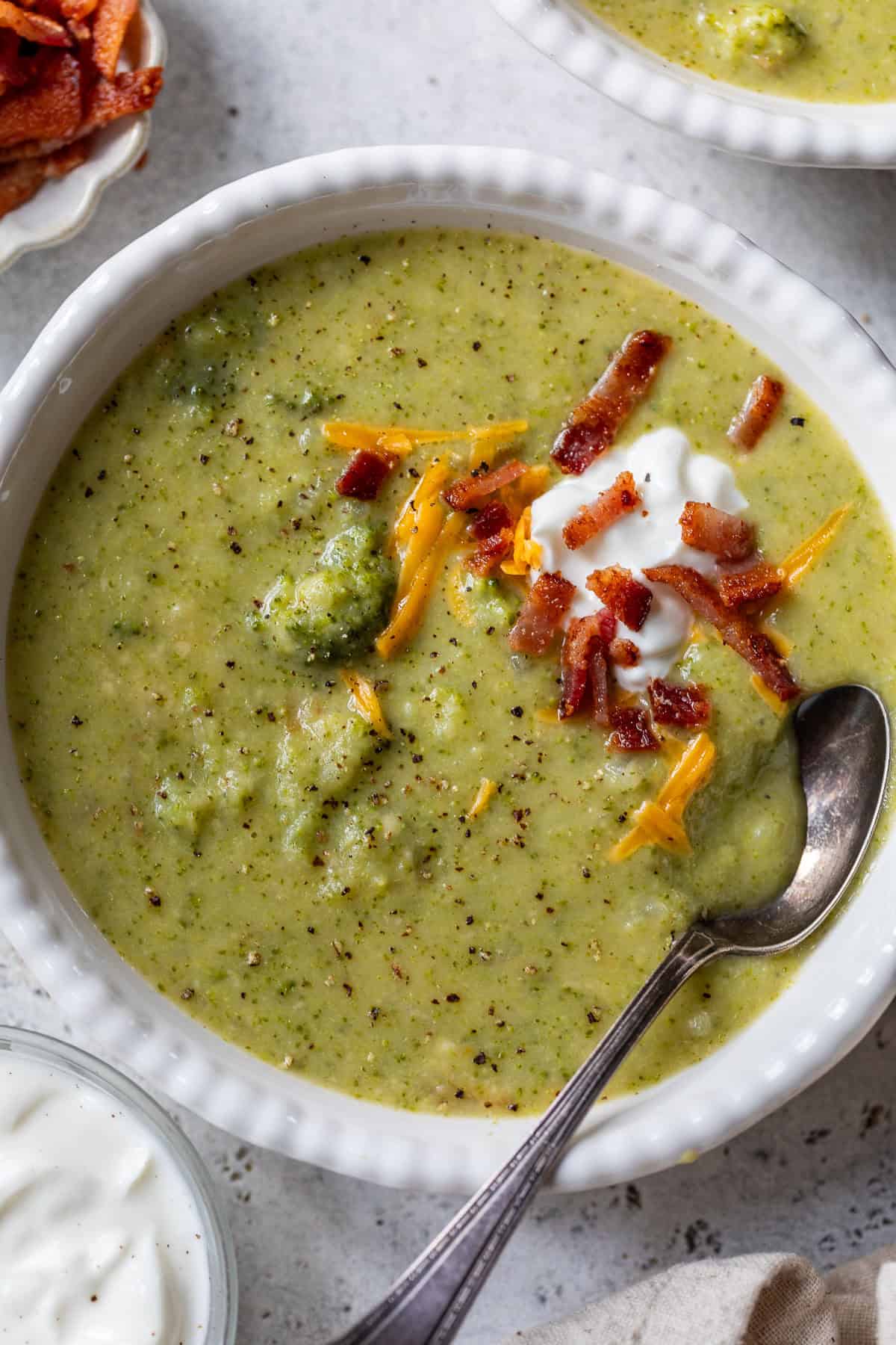 Creamy broccoli potato soup topped with shredded cheese, bacon bits, and a dollop of sour cream in a white bowl with a spoon.