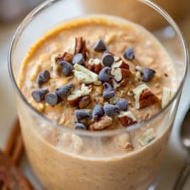 A glass of pumpkin overnight oats topped with chocolate chips and chopped pecans, with cinnamon sticks nearby.