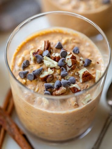 A glass of pumpkin overnight oats topped with chocolate chips and chopped pecans, with cinnamon sticks nearby.