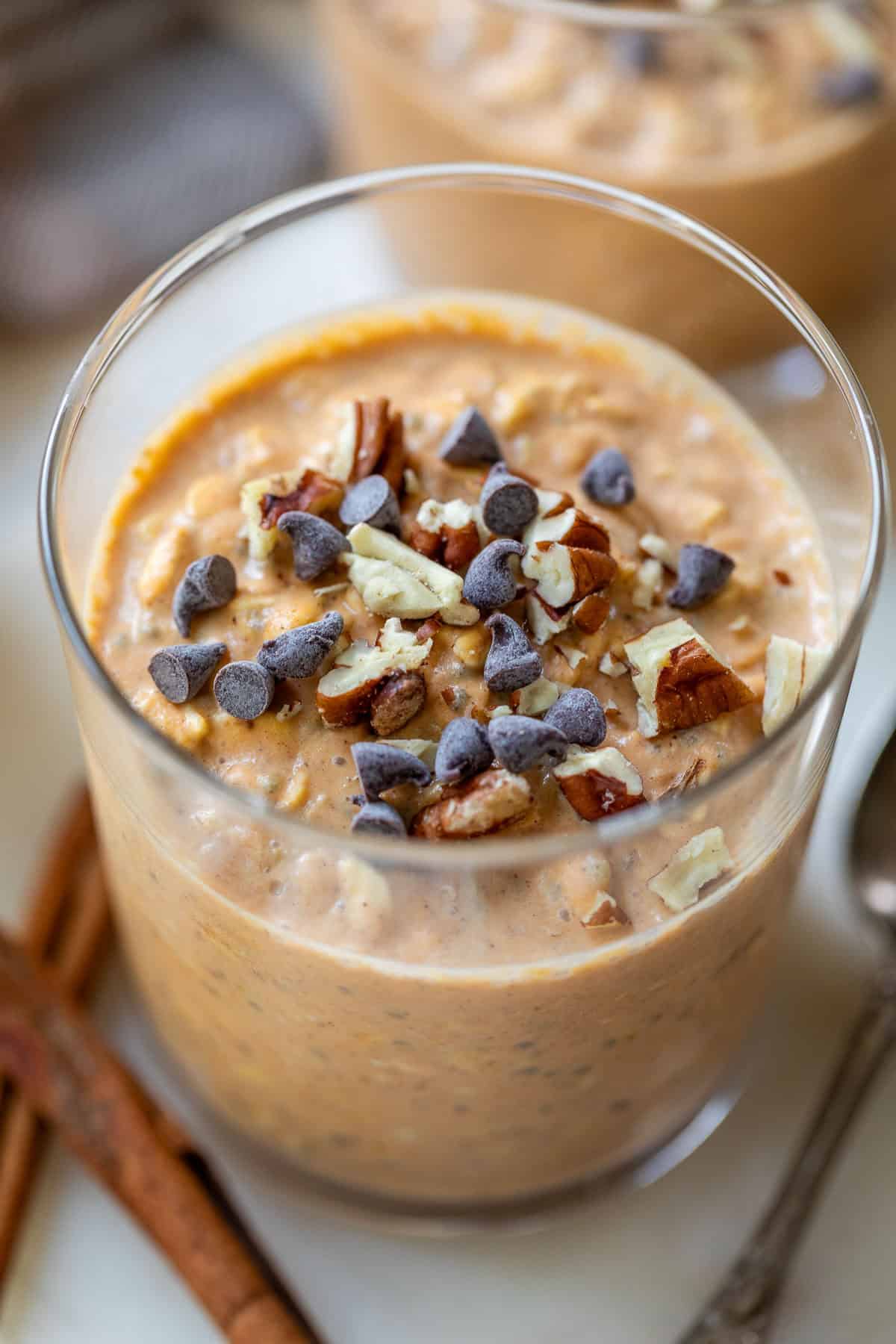 A glass of pumpkin overnight oats topped with chocolate chips and chopped pecans, with cinnamon sticks nearby.