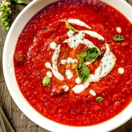 A bowl of roasted red pepper soup garnished with cream and fresh basil, on a wooden table with a spoon and basil leaves nearby.