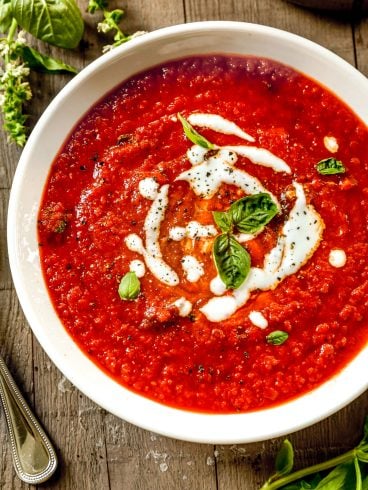 A bowl of roasted red pepper soup garnished with cream and fresh basil, on a wooden table with a spoon and basil leaves nearby.