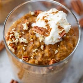 A glass of pumpkin oatmeal topped with whipped cream, chopped pecans, and a sprinkle of cinnamon.