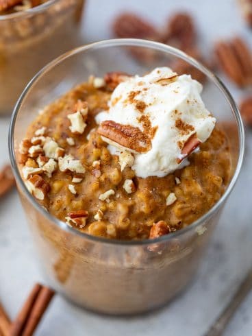 A glass of pumpkin oatmeal topped with whipped cream, chopped pecans, and a sprinkle of cinnamon.